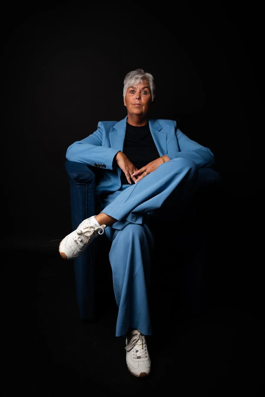 Arlette Vonk seated in an armchair, blue suit and white sneakers, looking straight at the camera