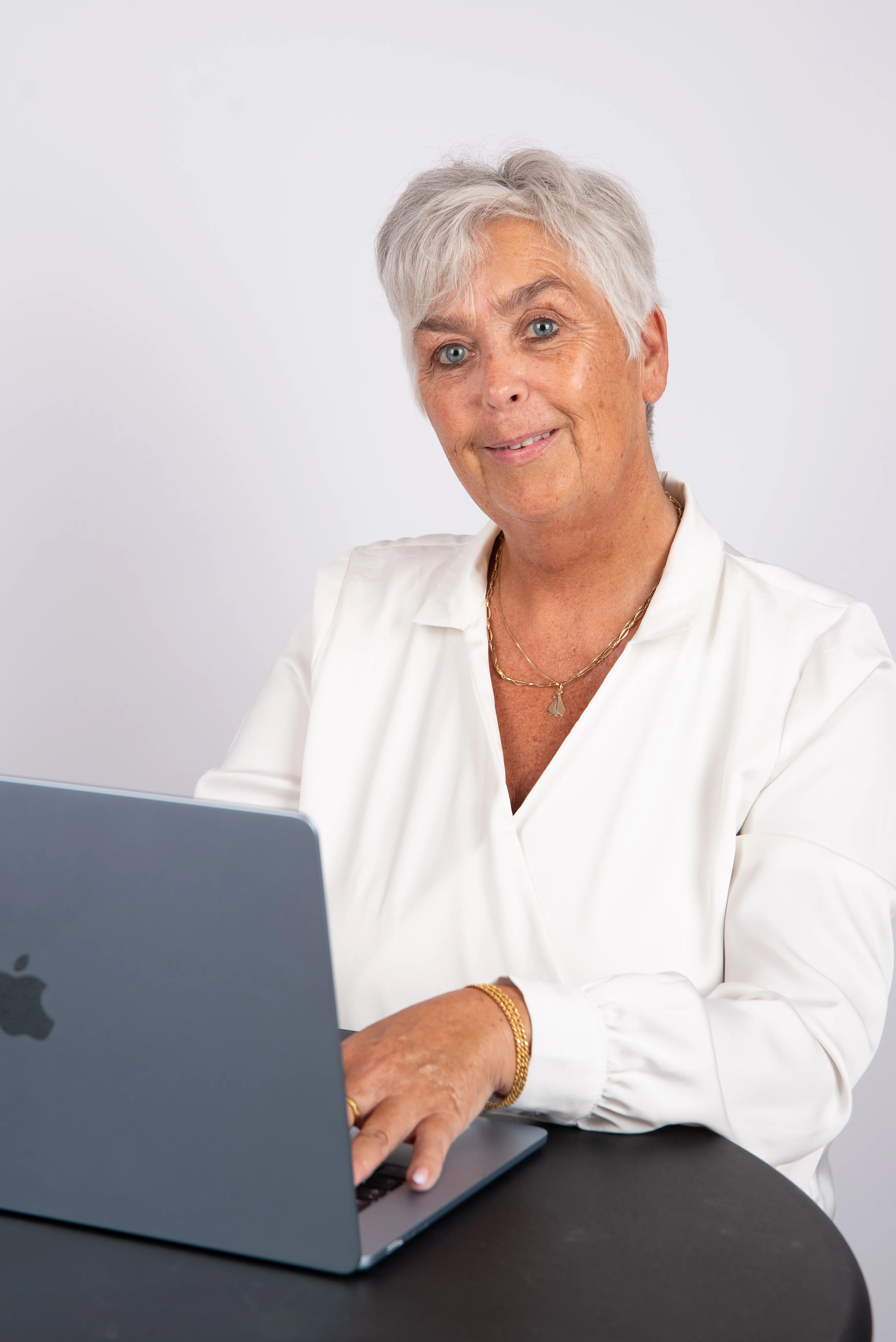 Arlette Vonk in a white blouse, working at a laptop at a round table against a pale backdrop, looking up at the camera