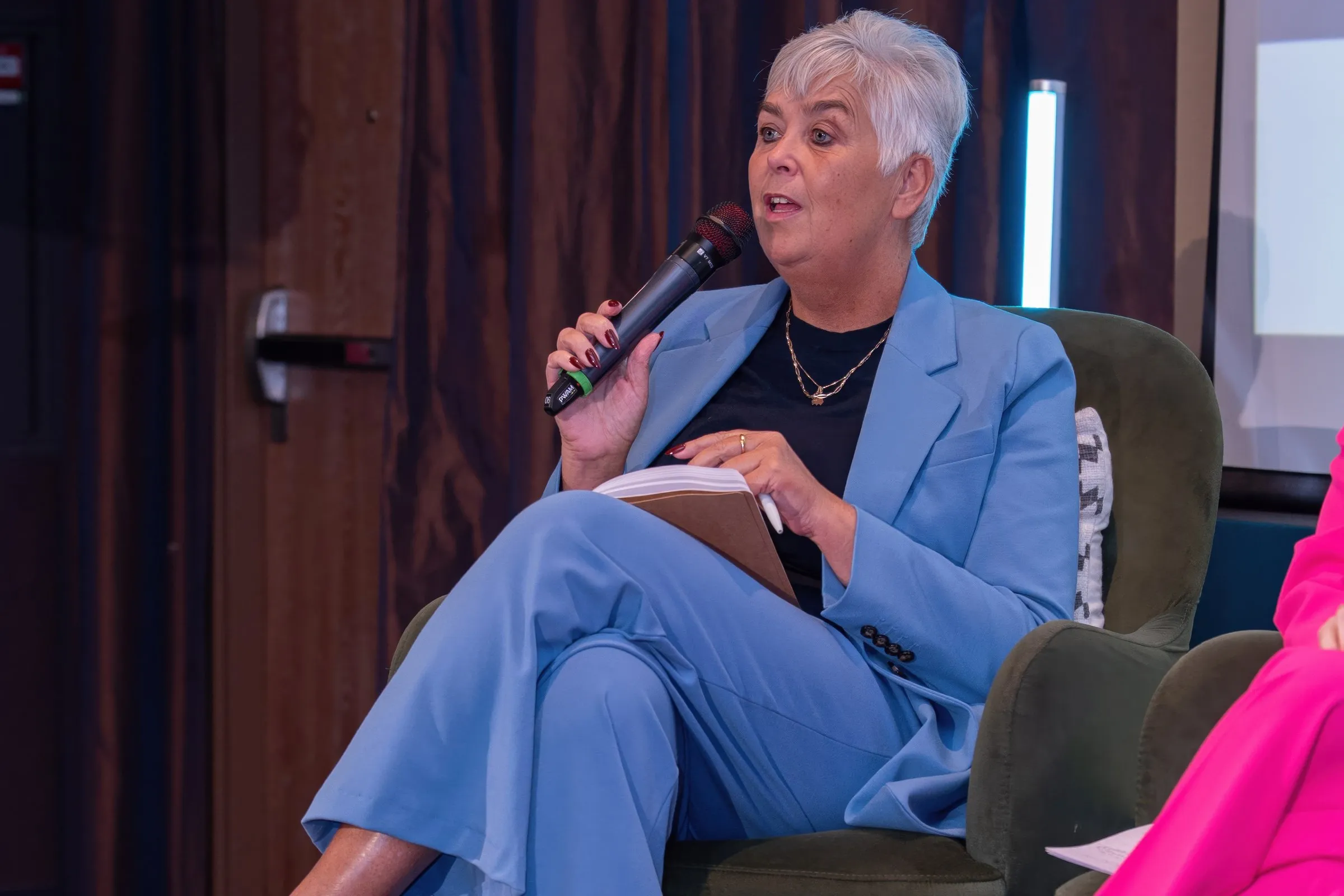 Arlette Vonk seated on a panel in a blue suit, speaking into a handheld microphone with a notebook on her lap